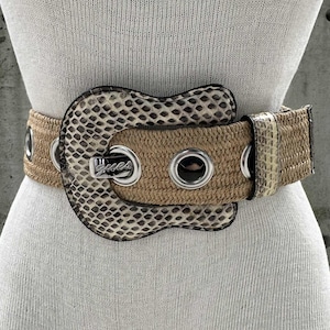 May include: A tan belt with a snakeskin-patterned buckle and accents. The belt features silver grommets and a silver buckle with the word "Guess". The belt is displayed on a mannequin.