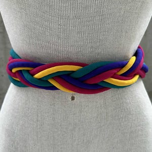 Może przedstawiać: Wielokolorowy pleciony pasek w odcieniach turkusu, fioletu, żółci i magenty. Pasek jest owinięty wokół manekina, prezentując jego pleciony wzór i żywą paletę barw. Ten dodatek dodaje koloru każdej stylizacji.