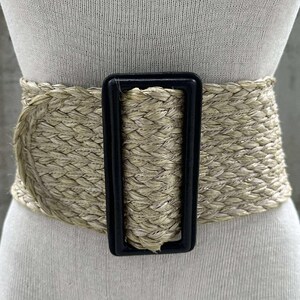 May include: A wide woven beige belt with a black buckle. The belt is made of a thick, braided material and has a simple, rectangular buckle.