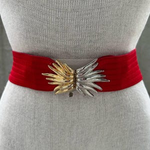 Puede incluir: Un cinturón de terciopelo rojo con una hebilla decorativa dorada y plateada. La hebilla tiene forma de flor estilizada con pétalos puntiagudos.