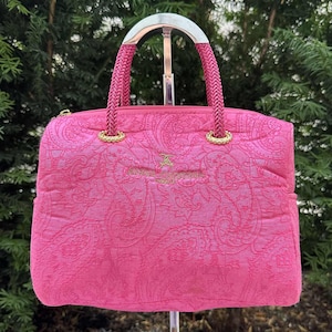 May include: A bright pink handbag with a paisley pattern. The bag has braided pink handles and gold-colored accents. The top handle is silver. The bag has a gold logo on the front.