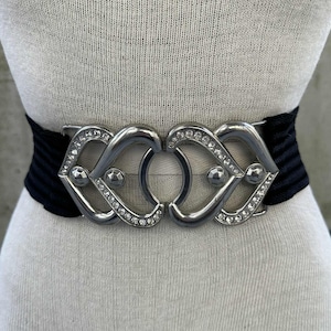 May include: Black elastic waist belt with a silver-tone buckle. The buckle features an interlocking design with clear rhinestones and decorative silver accents. The belt is displayed on a mannequin.
