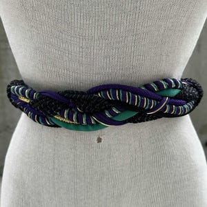 May include: Braided belt featuring a mix of black, purple, and teal colors. The belt has gold accents woven throughout the design. The belt is displayed on a mannequin, showcasing its woven texture and color combination.