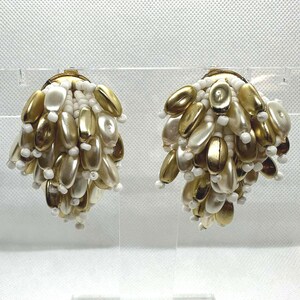 Może przedstawiać: Para klipsów z kolczykami o wzorze klastra. Kolczyki mają złotą metalową podstawę, z mieszanką złotych i kremowych owalnych koralików oraz małych białych koralików. Kolczyki są prezentowane na przezroczystym stojaku.