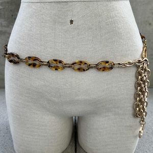 May include: A gold-toned chain belt with oval-shaped tortoise shell links. The belt is fastened around a mannequin and has a dangling chain detail. The belt is a vintage accessory.