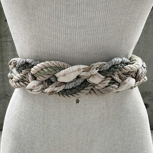 May include: Braided belt in neutral tones of beige, gray, and green. The belt is made of rope-like material and is displayed on a mannequin. The belt is a wide style, suitable for cinching the waist. The belt's design features a woven pattern.