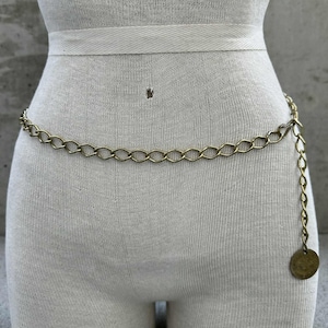 May include: A gold-colored chain belt with an oval link design. The belt is draped around a mannequin and features a dangling chain with a circular charm at the end. The belt is a vintage style.