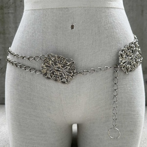 May include: Silver-toned metal belt with ornate, filigree-style rectangular and circular centerpieces. The belt features a double chain design and an adjustable chain with a ring closure. The belt is displayed on a mannequin.