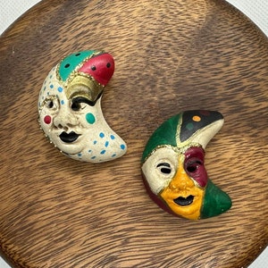 Może przedstawiać: Dwie ceramiczne maski w kształcie półksiężyca z namalowanymi twarzami. Jedna maska jest biała z zielonymi, czerwonymi i złotymi akcentami. Druga maska jest biała z zielonymi, czerwonymi, żółtymi i czarnymi akcentami.