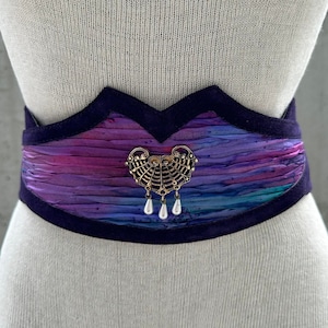 May include: A purple and blue patterned fabric belt with a gold metal ornament and pearl accents. The belt has a scalloped edge and is made of a soft, luxurious fabric.