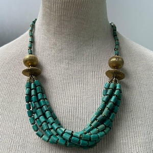 May include: A turquoise beaded necklace with multiple strands. The necklace features cylindrical beads and gold-colored accents. The design includes large, round gold beads at the top and a cascading arrangement of turquoise beads.