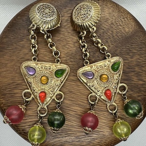 Może przedstawiać: Para złotych kolczyków wiszących z trójkątnym wisiorkiem ozdobionym kolorowymi kamieniami i koralikami. Kolczyki mają filigranowy wzór i są ozdobione zielonymi, czerwonymi, fioletowymi i żółtymi koralikami.