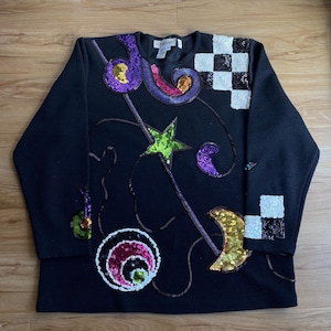 May include: Black long-sleeved sweater with colorful sequin embellishments in a geometric pattern. The design features stars, moons, and swirls. The sweater has a crew neck and is made of a soft, comfortable fabric.