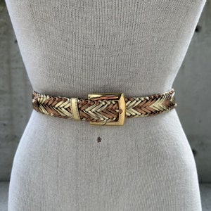 Może przedstawiać: Spleciony skórzany pasek z klamrą w kolorze złotym. Pasek jest wykonany z brązowej i złotej skóry.