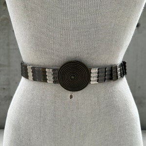 Może przedstawiać: Srebrny i brązowy pasek z koralików z dużą, okrągłą klamrą w kształcie sznura.