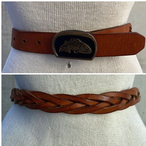 May include: Two brown leather belts with different buckle styles. The top belt has a large, ornate buckle with a fish design. The bottom belt is braided and has a simple buckle.