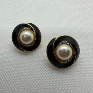 May include: A pair of black and gold earrings with a white pearl in the center of each earring. The earrings are round and have a swirl design.