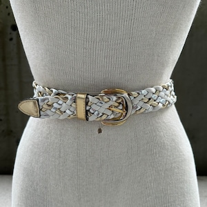 Może przedstawiać: Biały pleciony pasek ze złotą i srebrną klamrą. Pasek jest wykonany z połączenia białych, złotych i srebrnych nici.