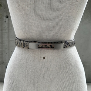 May include: A brown and black snakeskin belt with a silver buckle. The buckle is rectangular with a cut-out in the centre.