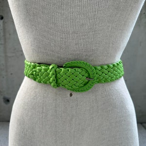 Puede incluir: Un cinturón tejido verde brillante con una hebilla plateada. El cinturón está hecho de un material trenzado y tiene un diseño clásico.