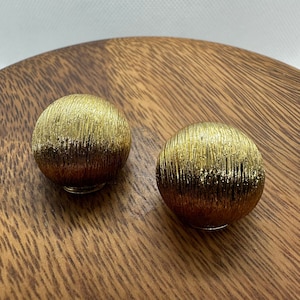 May include: A pair of gold-toned earrings with a textured, ridged surface. The earrings are round and have a slightly domed shape.