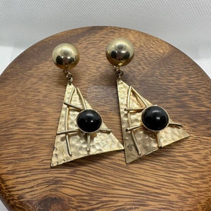 Może przedstawiać: Para złotych kolczyków na klipsie z czarnymi kamieniami kaboszonowymi osadzonymi w geometrycznym wzorze trójkąta.