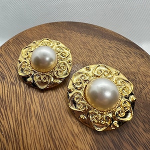 May include: A pair of gold-toned clip-on earrings with a large, pearlescent faux pearl center. The earrings have an ornate, scrolled design around the pearl, and are displayed on a wooden surface. The earrings are round and have a vintage aesthetic.