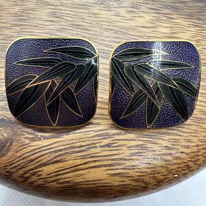 May include: A pair of square earrings with a gold trim. The earrings have a textured, dark purple background with a black and green bamboo leaf design. The earrings are displayed on a wooden surface.