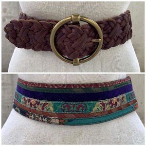 May include: Two belts, one brown leather with a braided design and a large circular buckle, and one multi-colored fabric belt with a purple velvet stripe.