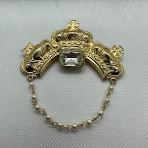 May include: A gold-tone brooch with a large clear rhinestone in the center. The brooch is shaped like three crowns with a pearl chain hanging from the bottom.