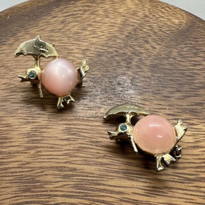 Puede incluir: Un par de broches doradas con una piedra cabujón rosa, un ojo de cristal verde y una sombrilla dorada.