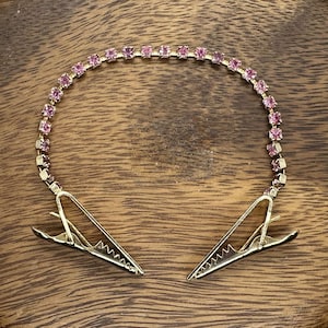May include: A pair of gold-tone hair clips with a pink rhinestone chain connecting them.