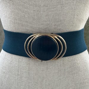 May include: A dark teal elastic belt featuring a central circular buckle with a textured dark teal disc surrounded by three concentric gold-toned metal rings. The belt is wide and designed to stretch, providing a secure fit around the waist.