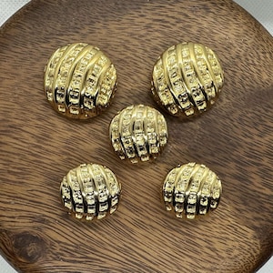 May include: Five gold-toned buttons with a textured, ribbed design. The buttons are arranged in a semi-circle on a wooden surface.