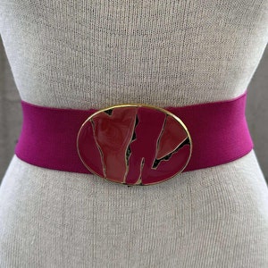 May include: A magenta elastic belt with a large oval buckle. The buckle has a gold-tone frame and a red and black abstract design. The belt is shown on a mannequin.