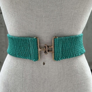 Może przedstawiać: Elastyczny pasek w kolorze turkusowym z  złotą klamrą. Pasek wykonany jest z rozciągliwego materiału i ma prosty design.