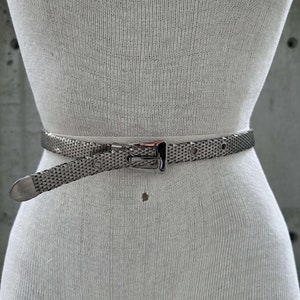 Peut inclure: Une ceinture en maille argent&eacute;e avec une boucle rectangulaire et une extr&eacute;mit&eacute; pointue. La ceinture est port&eacute;e autour de la taille d'un mannequin, mettant en valeur son design et sa texture. La ceinture comporte une s&eacute;rie de petits trous pour le r&eacute;glage.