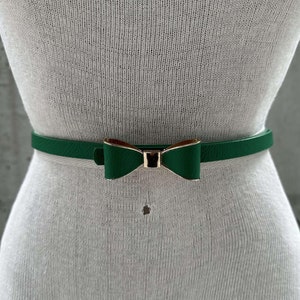 Puede incluir: Un cinturón de cuero verde con una hebilla de lazo dorada.