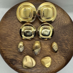 May include: A set of eight gold-tone earrings. The earrings are in various shapes, including round, oval, and square. The earrings are displayed on a wooden surface.