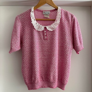 Może przedstawiać: Różowy sweter z krótkim rękawem, białym koronkowym kołnierzykiem i trzema białymi guzikami. Sweter ma fakturowany splot.