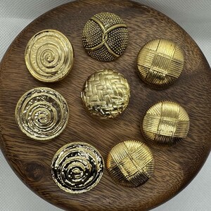 May include: Eight gold-toned buttons with different designs. The buttons are arranged in a circle on a wooden surface.