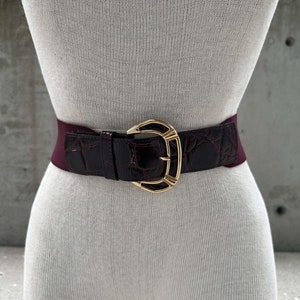 Puede incluir: Un cinturón elástico burdeos con una hebilla dorada con diseño de cocodrilo.