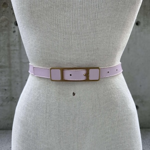 Puede incluir: Un cinturón de cuero rosa claro con una hebilla dorada. El cinturón se muestra en un maniquí blanco.