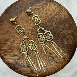 Może przedstawiać: Para złotych kolczyków wiszących z geometrycznymi wzorami i wieloma łańcuszkami z frędzlami.