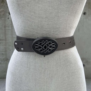 Może przedstawiać: Srebrny pasek skórzany w kolorze szarym z dużą, ozdobną klamrą w kształcie celtyckiego węzła. Klamra ma owalny kształt i fakturowane wykończenie.