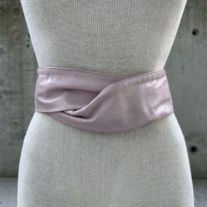 Puede incluir: Un cinturón de cuero rosa pálido con un diseño anudado. El cinturón está envuelto alrededor de un maniquí blanco.