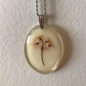 Może przedstawiać: Delikatny, biały kwiat wciśnięty w przezroczysty, owalny wisiorek z żywicy, wiszący na srebrnym łańcuszku.