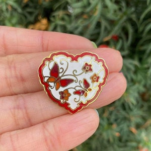 Puede incluir: Broche en forma de corazón con centro blanco, borde rojo y ribete dorado. El broche presenta mariposas rojas y naranjas y pequeñas flores. El fondo es un follaje verde borroso.