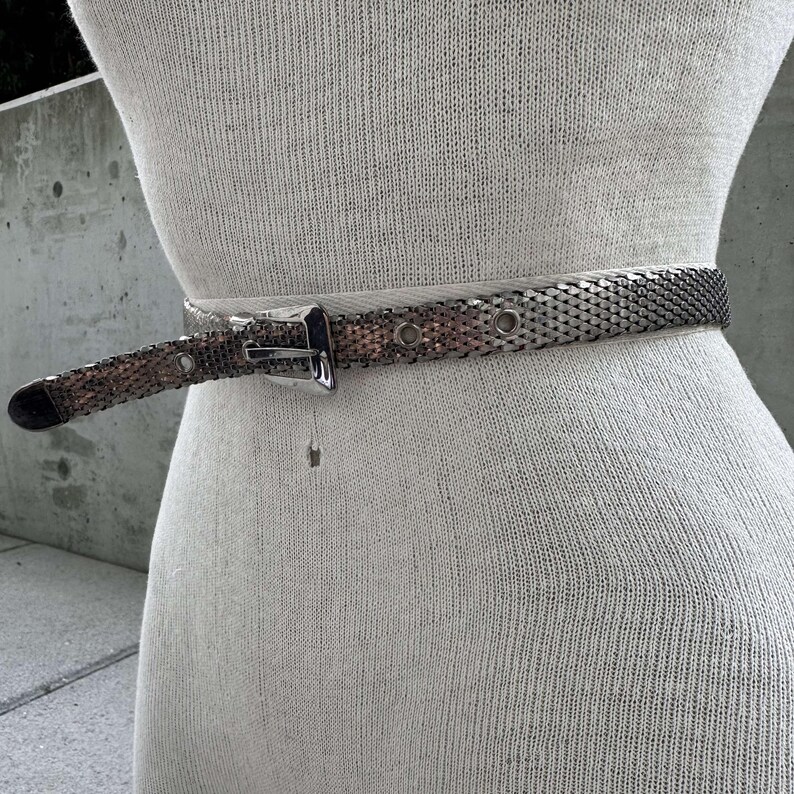 Peut inclure: Une ceinture en maille argent&eacute;e avec une boucle rectangulaire argent&eacute;e est pr&eacute;sent&eacute;e sur un mannequin. La ceinture a un design tiss&eacute; et est attach&eacute;e autour de la taille du mannequin. L'extr&eacute;mit&eacute; de la ceinture est l&eacute;g&egrave;rement plus fonc&eacute;e.