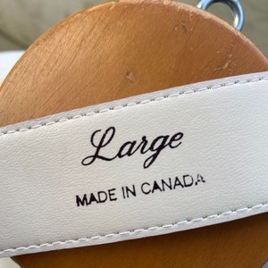 May include: Close-up of a white leather belt with the word "Large" in cursive script. The text "MADE IN CANADA" is printed below. The belt is displayed on a brown wooden form.
